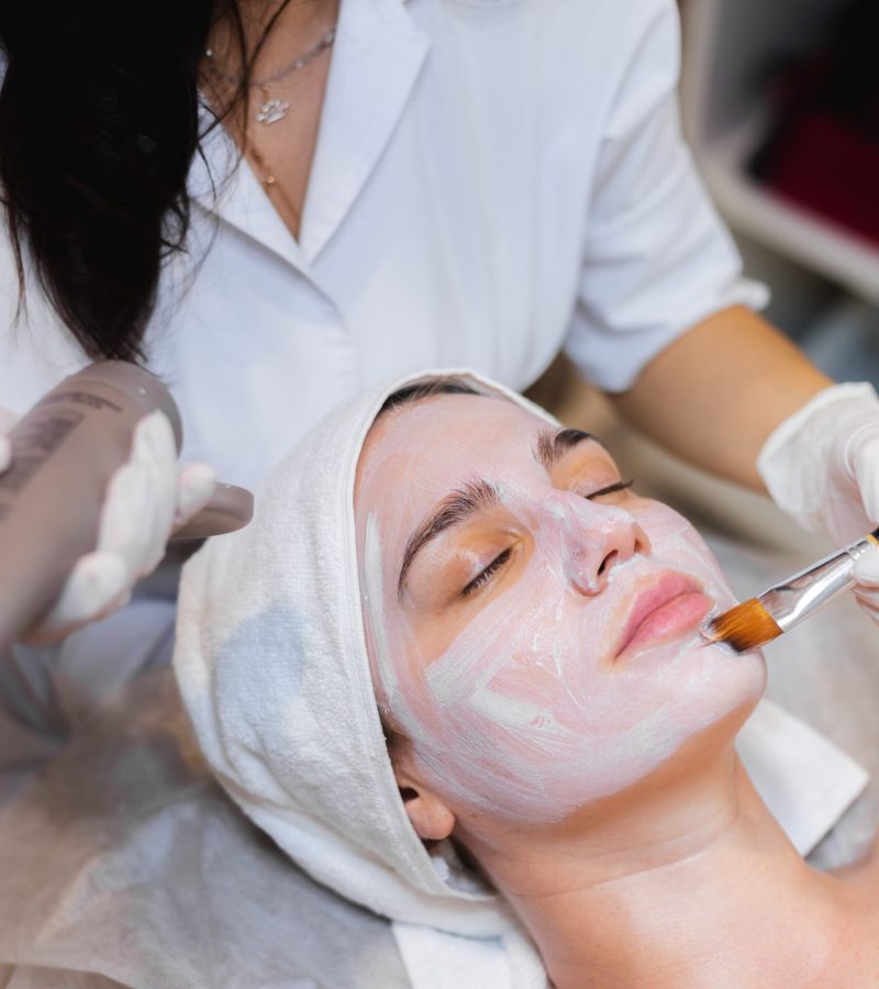 Beautician with a brush applies a white moisturizing mask to the face of a young girl client in a spa beauty salon